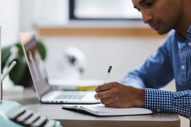 Pessoa escrevendo metas financeiras em caderno ao lado do computador